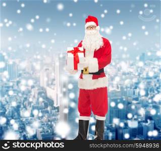 christmas, holidays and people concept - man in costume of santa claus with gift box over snowy city background