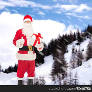 christmas, holidays and people concept - man in costume of santa claus with gift box showing thumbs up gesture over snowy mountains background