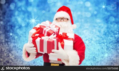 christmas, holidays and people concept - man in costume of santa claus with gift boxes over blue holidays lights background