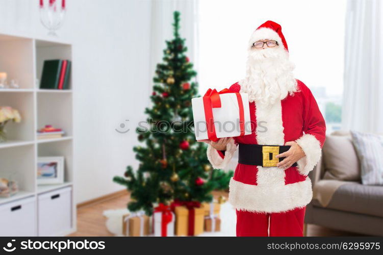 christmas, holidays and people concept - man in costume of santa claus with gift box over home room background. santa claus with christmas gift at home