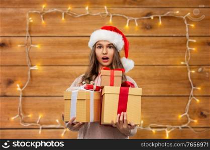 christmas, holidays and people concept - shocked teenage girl in santa helper hat holding gift box over garland lights on wooden background. teenage girl in santa hat with christmas gift