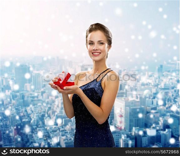 christmas, holidays and people concept - smiling woman in evening dress with small red gift box over snowy city background