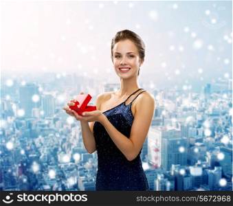 christmas, holidays and people concept - smiling woman in evening dress with small red gift box over snowy city background