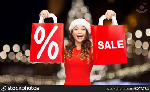 christmas, holidays and people concept - smiling young woman in santa hat with shopping bags with percent and sale sign over night lights background. happy woman insanta hat with shopping bags