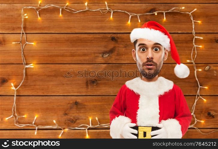christmas, holidays and people concept - surprised man in costume of santa over garland on wooden background (funny cartoon style character with big head). surprised santa over garland on wooden background