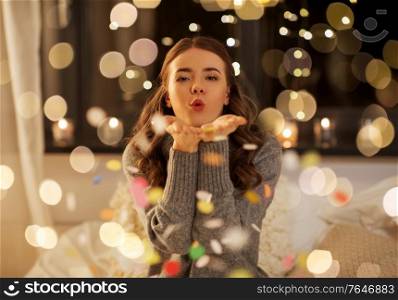 christmas, holidays and people concept - young woman blowing colorful confetti from her hands at home. woman blowing confetti from her hands at night