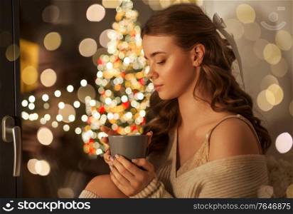 christmas, holidays and people concept - young woman in pullover sitting at window with coffee or tea cup at home. woman with coffee at window on christmas