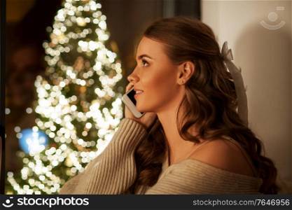christmas, holidays and people concept - young woman in pullover sitting at window at home and calling on smartphone at night. woman calling on smartphone at christmas night