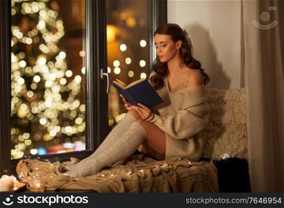 christmas, holidays and people concept - young woman in pullover sitting on windowsill and reading book at home. woman reading book on windowsill on christmas