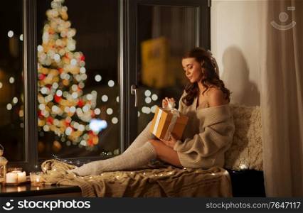 christmas, holidays and people concept - young woman in pullover sitting on windowsill with gift box at home at night. woman with christmas gift at home at night