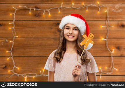 christmas, holidays and photo booth concept - happy smiling teenage girl in santa helper hat with gingerbread man party accessory over garland lights on wooden background. happy teenage girl in santa hat on christmas