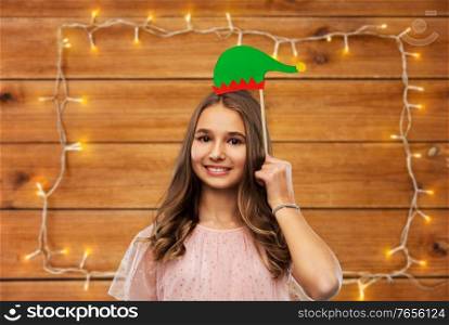 christmas, holidays and photo booth concept - happy smiling teenage girl with santa helper hat party accessory over garland lights on wooden background. happy teenage girl with santa helper hat accessory
