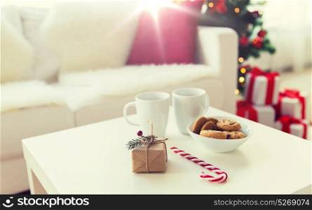 christmas, holidays and winter concept - close up of gift, oat cookies, sugar cane candy and cups on table at home. close up of gift, sweets and cups on table at home