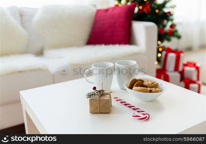 christmas, holidays and winter concept - close up of gift, oat cookies, sugar cane candy and cups on table at home