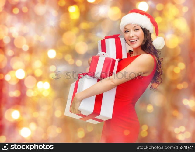 christmas, holidays, celebration and people concept - happy smiling woman in santa hat and red dress holding gift boxes over lights background