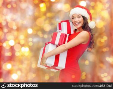 christmas, holidays, celebration and people concept - happy smiling woman in santa hat and red dress holding gift boxes over lights background