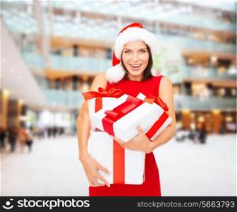 christmas, holidays, celebration and people concept - smiling woman in santa helper hat and red dress with gift boxes over shopping center background