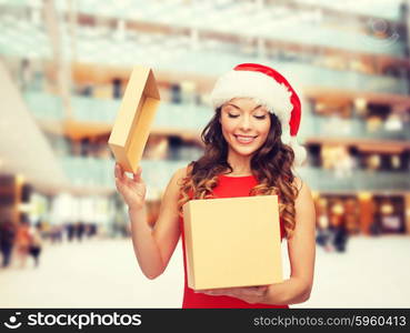 christmas, holidays, celebration and people concept - smiling woman in santa helper hat with gift box over shopping center background