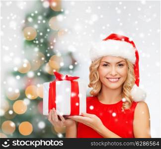 christmas, holidays, celebration and people concept - smiling woman in santa helper hat and red dress with gift box over tree lights background