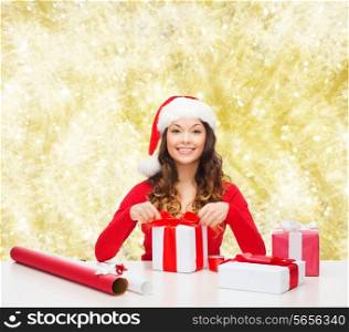 christmas, holidays, celebration, decoration and people concept - smiling woman in santa helper hat with decorating paper packing gift boxes over yellow lights background