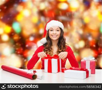 christmas, holidays, celebration, decoration and people concept - smiling woman in santa helper hat with decorating paper packing gift boxes over red lights background