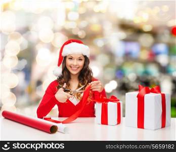 christmas, holidays, celebration, decoration and people concept - smiling woman in santa helper hat with scissors packing gift box over lights background
