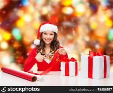 christmas, holidays, celebration, decoration and people concept - smiling woman in santa helper hat with scissors packing gift box over red lights background