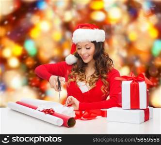 christmas, holidays, celebration, decoration and people concept - smiling woman in santa helper hat with scissors packing gift box over red lights background