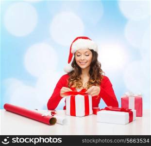 christmas, holidays, celebration, decoration and people concept - smiling woman in santa helper hat with decorating paper packing gift boxes over blue lights background