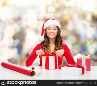 christmas, holidays, celebration, decoration and people concept - smiling woman in santa helper hat with decorating paper packing gift boxes over lights background