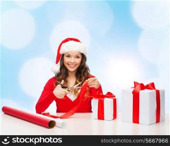 christmas, holidays, celebration, decoration and people concept - smiling woman in santa helper hat with scissors packing gift box over blue lights background