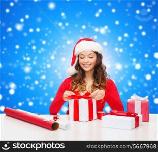 christmas, holidays, celebration, decoration and people concept - smiling woman in santa helper hat with decorating paper packing gift boxes over blue snowy background