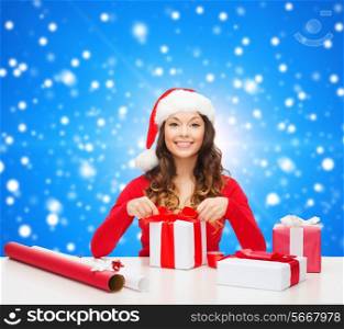 christmas, holidays, celebration, decoration and people concept - smiling woman in santa helper hat with decorating paper packing gift boxes over blue snowing background
