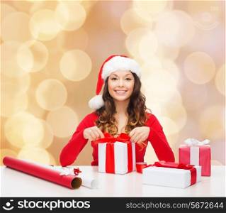 christmas, holidays, celebration, decoration and people concept - smiling woman in santa helper hat with decorating paper packing gift boxes over beige lights background