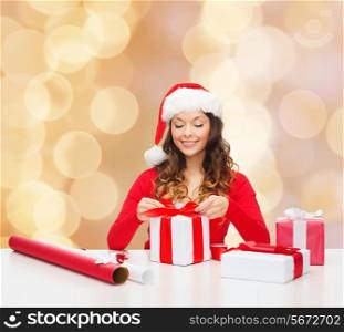 christmas, holidays, celebration, decoration and people concept - smiling woman in santa helper hat with decorating paper packing gift boxes over beige lights background