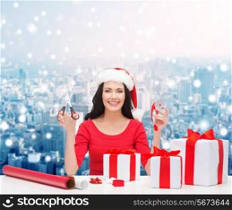 christmas, holidays, celebration, decoration and people concept - smiling woman in santa helper hat with scissors packing gift boxes over snowy city background