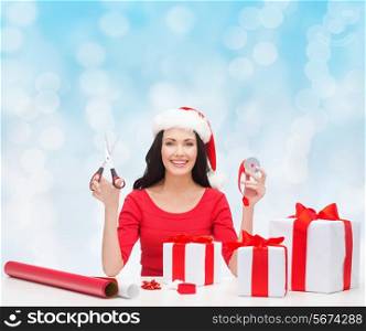 christmas, holidays, celebration, decoration and people concept - smiling woman in santa helper hat with scissors packing gift boxes over blue lights background