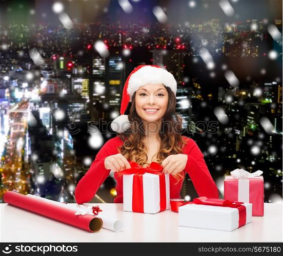 christmas, holidays, celebration, decoration and people concept - smiling woman in santa helper hat with decorating paper packing gift boxes over snowy night city background