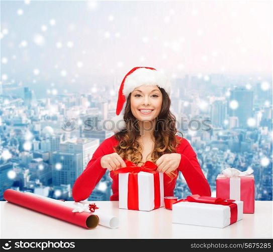christmas, holidays, celebration, decoration and people concept - smiling woman in santa helper hat with decorating paper packing gift boxes over snowy city background