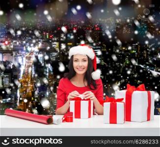 christmas, holidays, celebration, decoration and people concept - smiling woman in santa helper hat with decorating paper packing gift boxes over snowy night city background