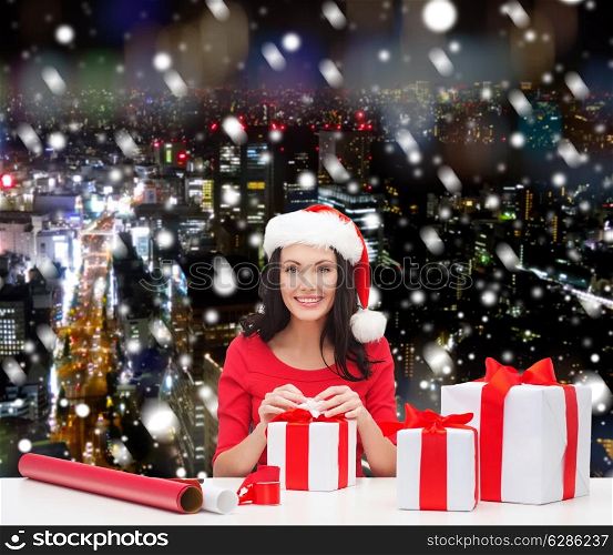 christmas, holidays, celebration, decoration and people concept - smiling woman in santa helper hats with decorating paper and gift boxes over snowy night city background