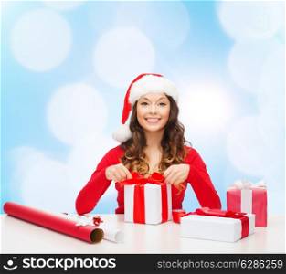 christmas, holidays, celebration, decoration and people concept - smiling woman in santa helper hat with decorating paper packing gift boxes over blue lights background