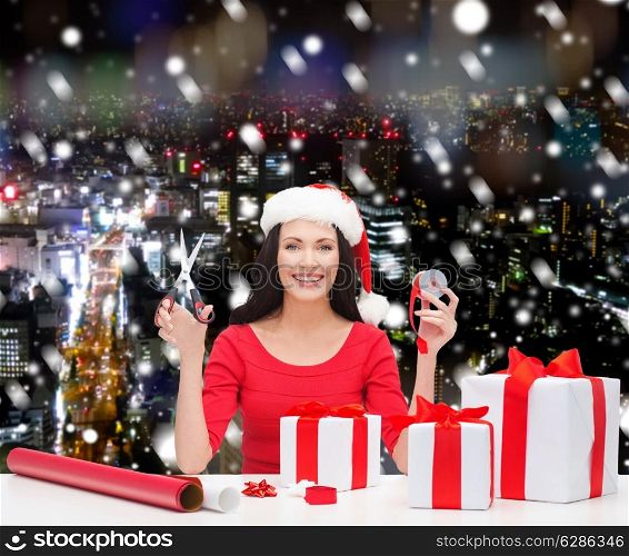 christmas, holidays, celebration, decoration and people concept - smiling woman in santa helper hat with scissors packing gift boxes over snowy night city background