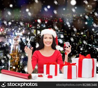 christmas, holidays, celebration, decoration and people concept - smiling woman in santa helper hat with scissors packing gift boxes over snowy night city background