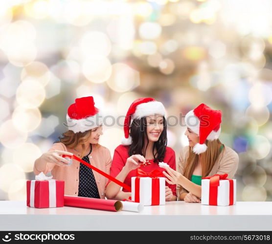 christmas, holidays, celebration, decoration and people concept - smiling women in santa helper hats with decorating paper and gift boxes over lights background