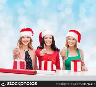 christmas, holidays, celebration, decoration and people concept - smiling women in santa helper hats with decorating paper and gift boxes showing thumbs up over blue lights background