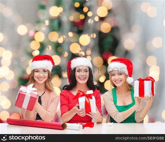 christmas, holidays, celebration, decoration and people concept - smiling women in santa helper hats with decorating paper and gift boxes over tree lights background