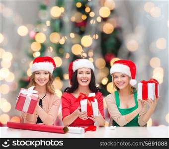 christmas, holidays, celebration, decoration and people concept - smiling women in santa helper hats with decorating paper and gift boxes over tree lights background