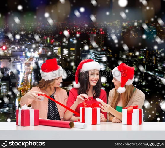 christmas, holidays, celebration, decoration and people concept - smiling women in santa helper hats with decorating paper and gift boxes over snowy night city background