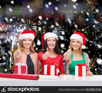 christmas, holidays, celebration, decoration and people concept - smiling women in santa helper hats with decorating paper and gift boxes showing thumbs up over snowy night city background